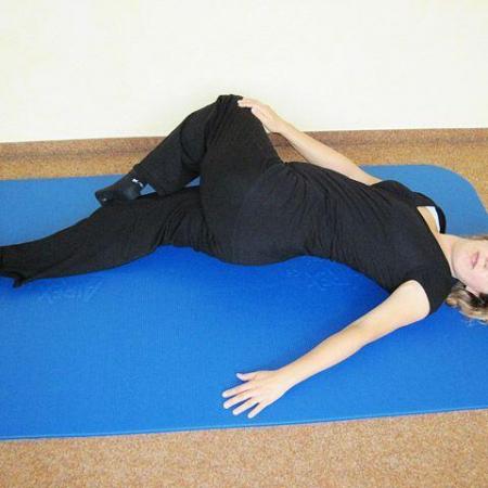 Schwangere Frau liegt in Rückenlage auf einer Yogamatte, das rechte Bein ist angewinkelt über das linke Bein gelegt, der rechte Arm liegt ausgestreckt zur Seite für eine sanfte Dehnung der Wirbelsäule.