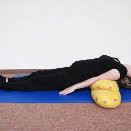 Schwangere Frau liegt entspannt in Rückenlage auf einer blauen Yogamatte, der Oberkörper wird von einem gelben Kissen gestützt, Arme ruhen locker neben dem Körper.