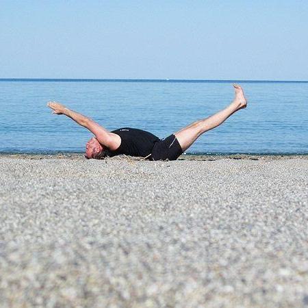 Ein älterer Mann in schwarzer Sportkleidung liegt barfuß auf dem Rücken auf einem kiesigen Strand vor ruhigem Meer. Er hebt gleichzeitig ein gestrecktes Bein und beide Arme vom Boden, der Blick geht nach oben. Im Hintergrund ist eine klare Wasserlinie unter blauem Himmel zu sehen.